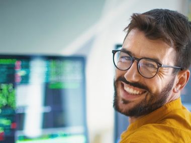 Lächelnder Mann mit Brille an einem Arbeitsplatz mit mehreren Monitoren.